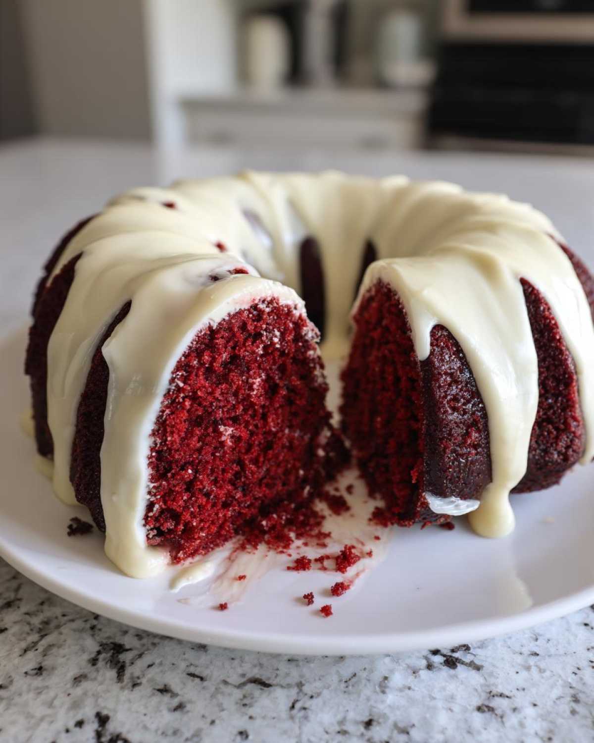 A moist red velvet bundt cake with a thick, white cream cheese glaze dripping down the sides, sitting on a white plate.