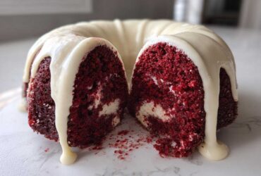 A moist red velvet bundt cake, cut in half to show the interior texture and cream cheese swirl, topped with dripping white glaze.