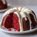 Close-up of a moist red velvet bundt cake with a thick, white cream cheese glaze dripping down the sides, a slice removed.