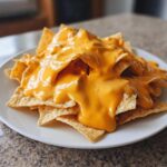 Close-up of tortilla chips smothered in thick, bright orange nacho cheese sauce on a white plate.
