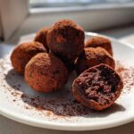 A pile of rich, dark chocolate protein balls dusted heavily with cocoa powder on a white plate.
