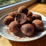 A plate of rich, dark chocolate protein balls dusted heavily with cocoa powder, one cut open.