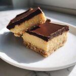 Two squares of no-bake peanut butter bars with a thick chocolate ganache layer on a white plate.