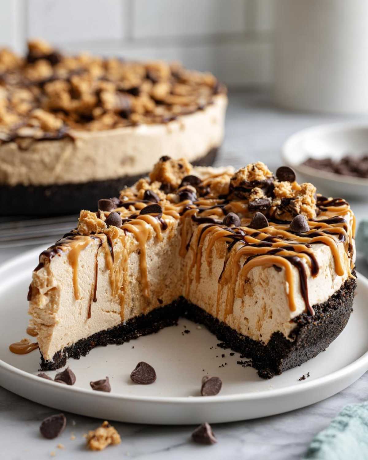 A slice removed from a creamy no-bake peanut butter pie, drizzled with caramel and chocolate, topped with cookie crumbles.