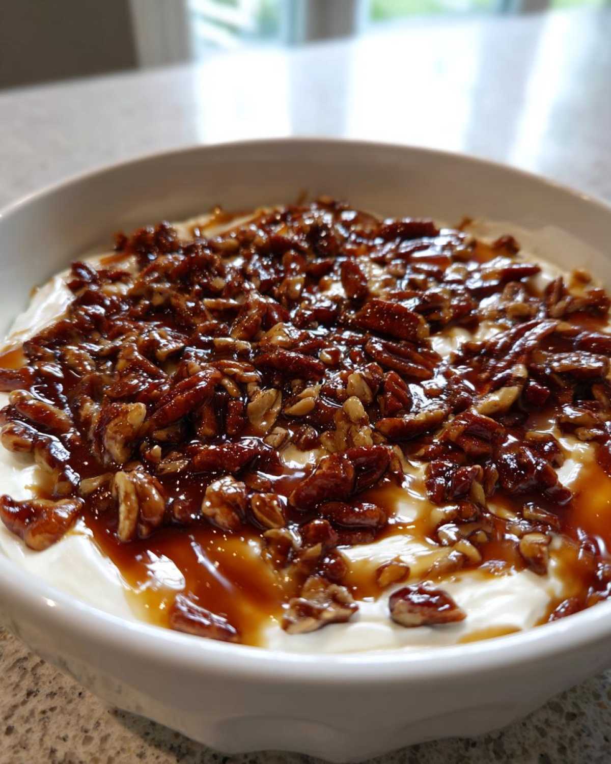 A close-up of a white bowl filled with creamy no-bake pecan pie dip, topped generously with candied pecans and caramel sauce.