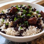 A close-up of a white bowl filled with fluffy white rice topped generously with seasoned black beans and chunks of savory meat, garnished with fresh parsley.