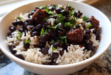 A close-up of a white bowl filled with fluffy white rice topped generously with seasoned black beans and chunks of savory meat, garnished with fresh parsley.