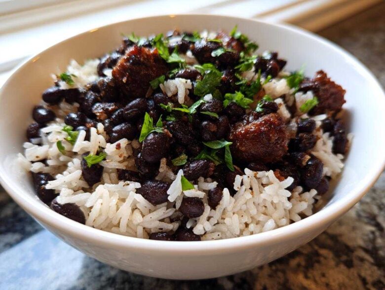 A close-up of a white bowl filled with fluffy white rice topped generously with seasoned black beans and chunks of savory meat, garnished with fresh parsley.