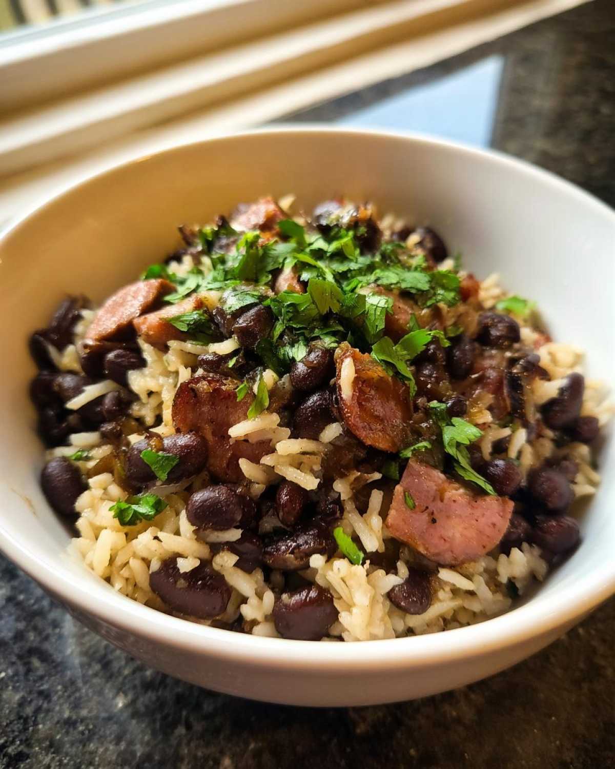 Close-up of a white bowl filled with savory black beans and rice mixed with slices of smoked sausage and topped with fresh cilantro.