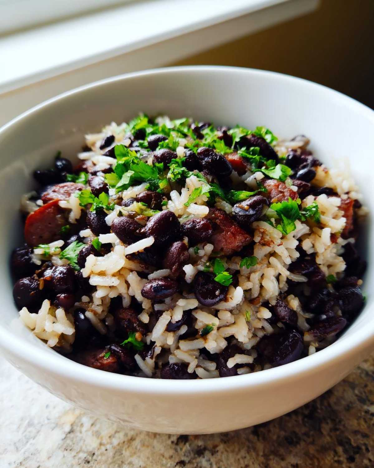 Close-up of a white bowl filled with flavorful black beans and rice mixed with pieces of sausage and topped with fresh cilantro.