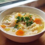 A close-up of a white bowl filled with hot penicillin soup, featuring shredded chicken, pasta noodles, carrots, and fresh parsley.