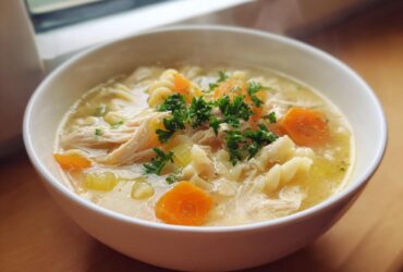 A close-up of a white bowl filled with hot penicillin soup, featuring shredded chicken, pasta noodles, carrots, and fresh parsley.