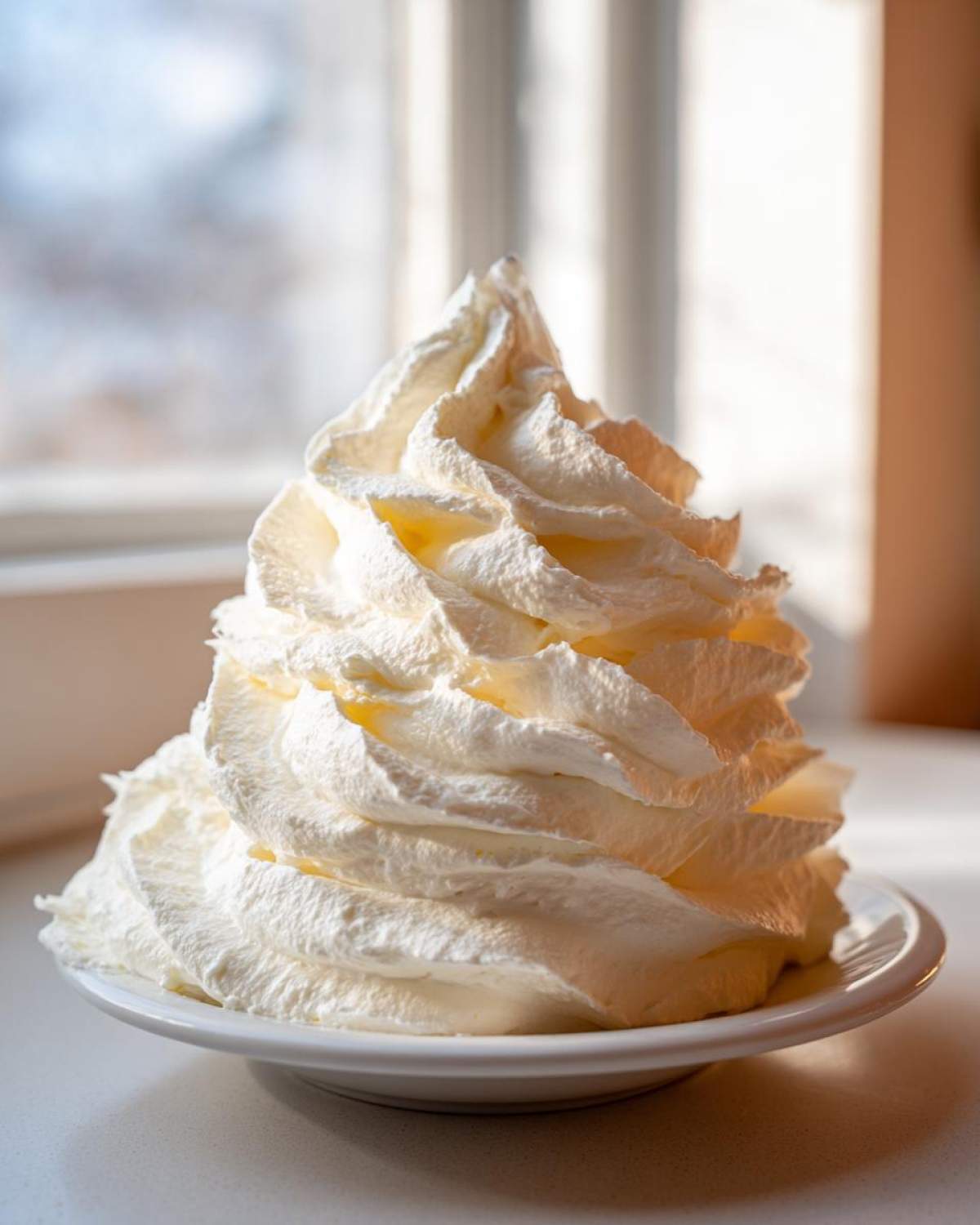 Tall swirl of fluffy, white cream cheese frosting piped high on a small white plate, backlit by a window.