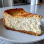 A thick slice of creamy Protein Cheesecake with a graham cracker crust and a lightly browned top, served on a white plate.