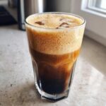 A tall glass filled with dark cold brew coffee topped with thick, light brown foam, perfect for a pumpkin cream cold brew.