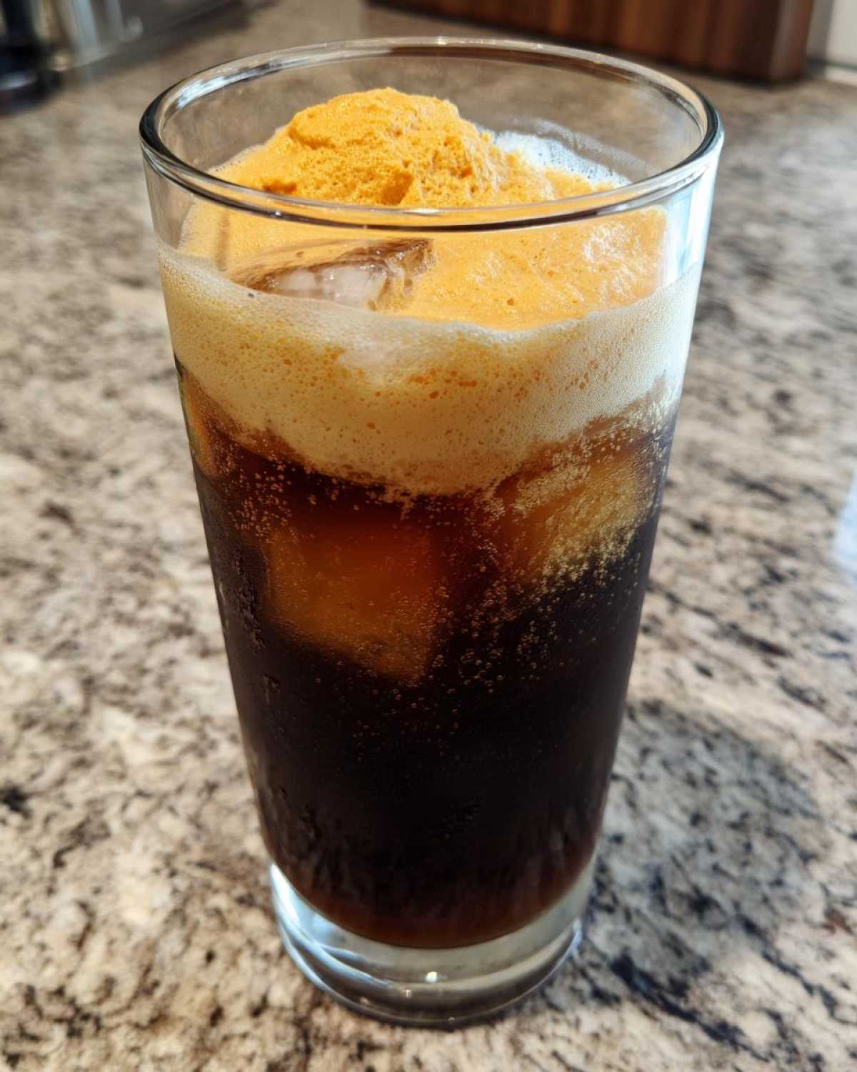 Close-up of a tall glass filled with pumpkin cream cold brew, showing dark coffee, ice, and thick orange foam.
