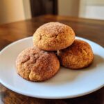 Three soft, round pumpkin snickerdoodle cookies stacked on a white plate, coated in cinnamon sugar.