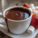 A white cup filled with rich chocolate fondue, served alongside fresh strawberries and marshmallows for dipping.