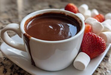 A white cup filled with rich chocolate fondue, served alongside fresh strawberries and marshmallows for dipping.