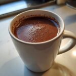 A close-up of rich, dark homemade hot cocoa with foam resting in a white ceramic mug.