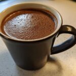 Close-up of a dark mug filled with frothy homemade hot cocoa resting on a light countertop.