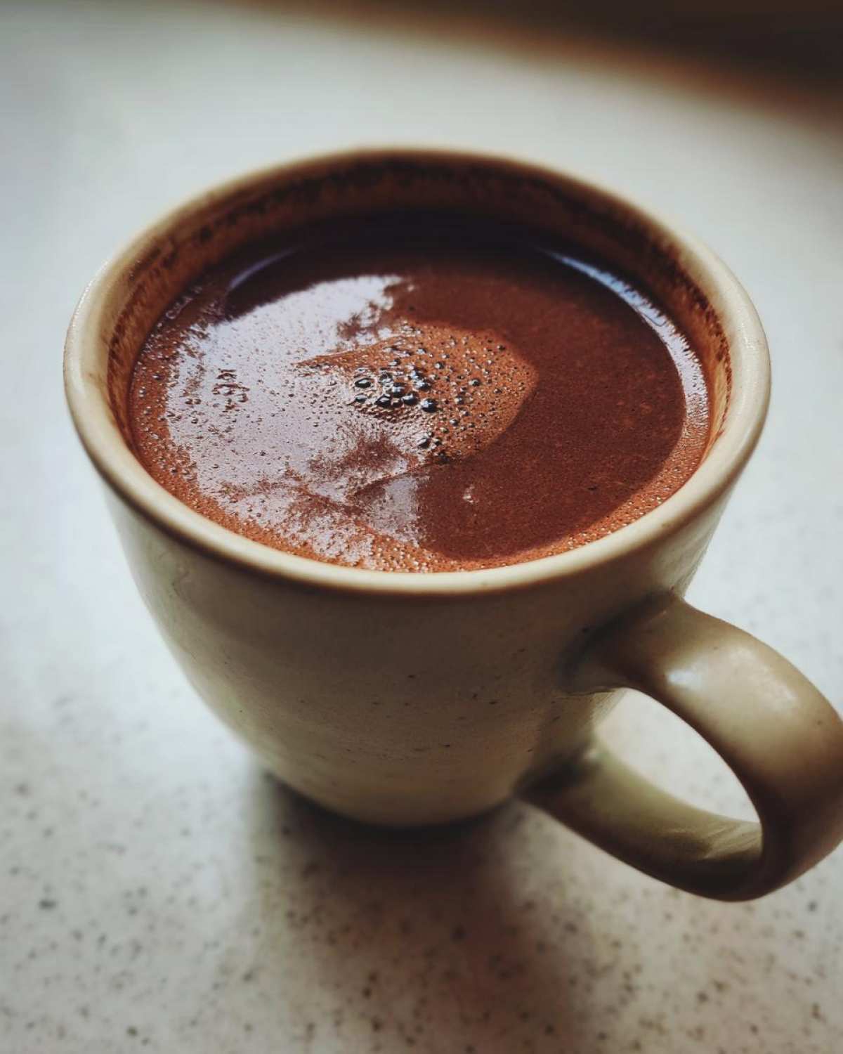 Close-up of rich, dark homemade hot cocoa with a slight foam layer in a speckled ceramic mug.