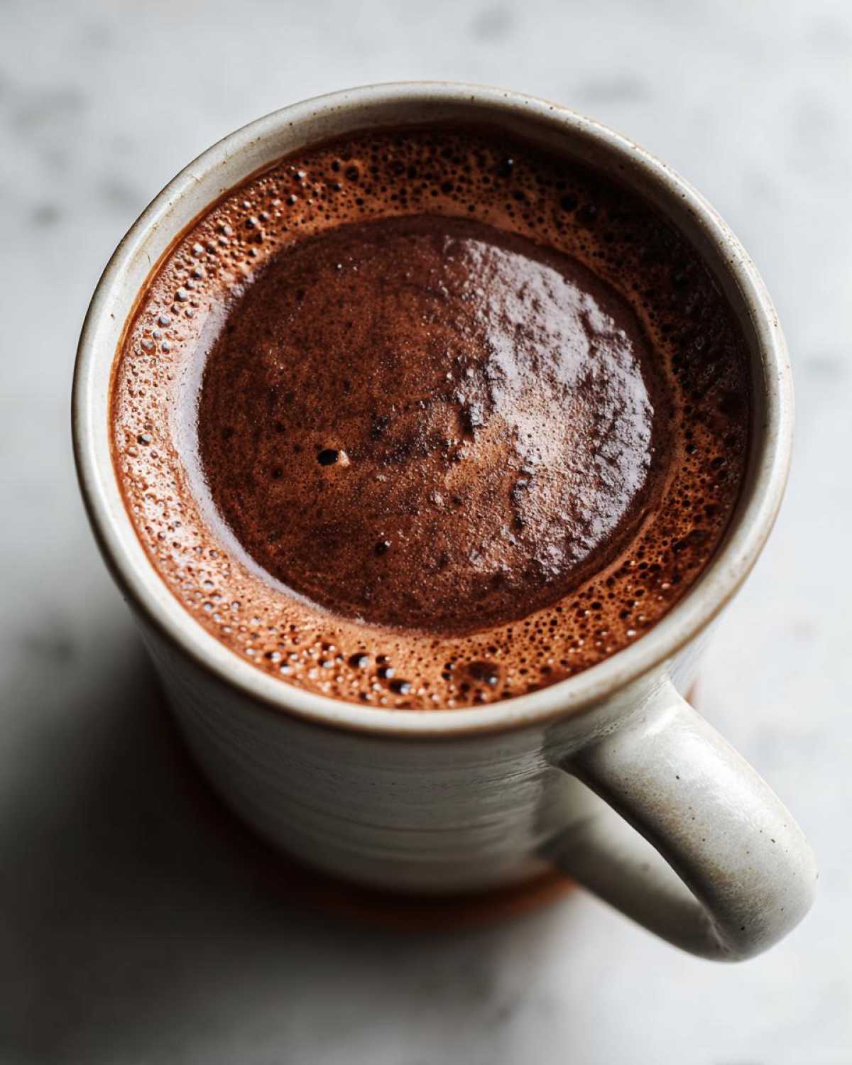 Overhead view of rich, dark homemade hot cocoa with a foamy top in a textured ceramic mug.