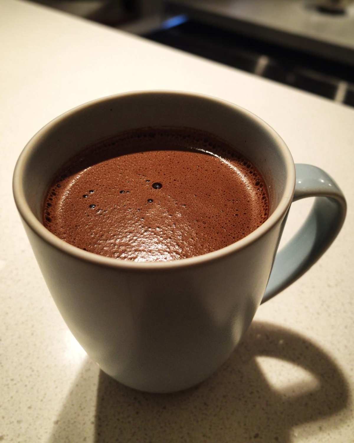 Close-up of rich, foamy homemade hot cocoa in a light blue ceramic mug sitting on a counter.