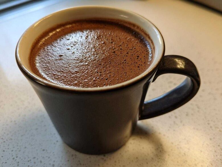 Close-up of a dark mug filled with frothy homemade hot cocoa resting on a light countertop.