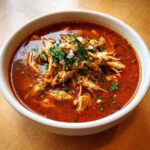 A close-up of a white bowl filled with vibrant red chicken pozole, topped with shredded chicken, cilantro, and cotija cheese.