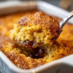 Close-up of a spoonful of warm, golden-brown apple cobbler being lifted from a white baking dish, showing the moist cake and syrupy filling.
