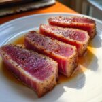 Four thick slices of perfectly seared tuna steak, showing a rare pink center and seasoned crust, resting in a light marinade.
