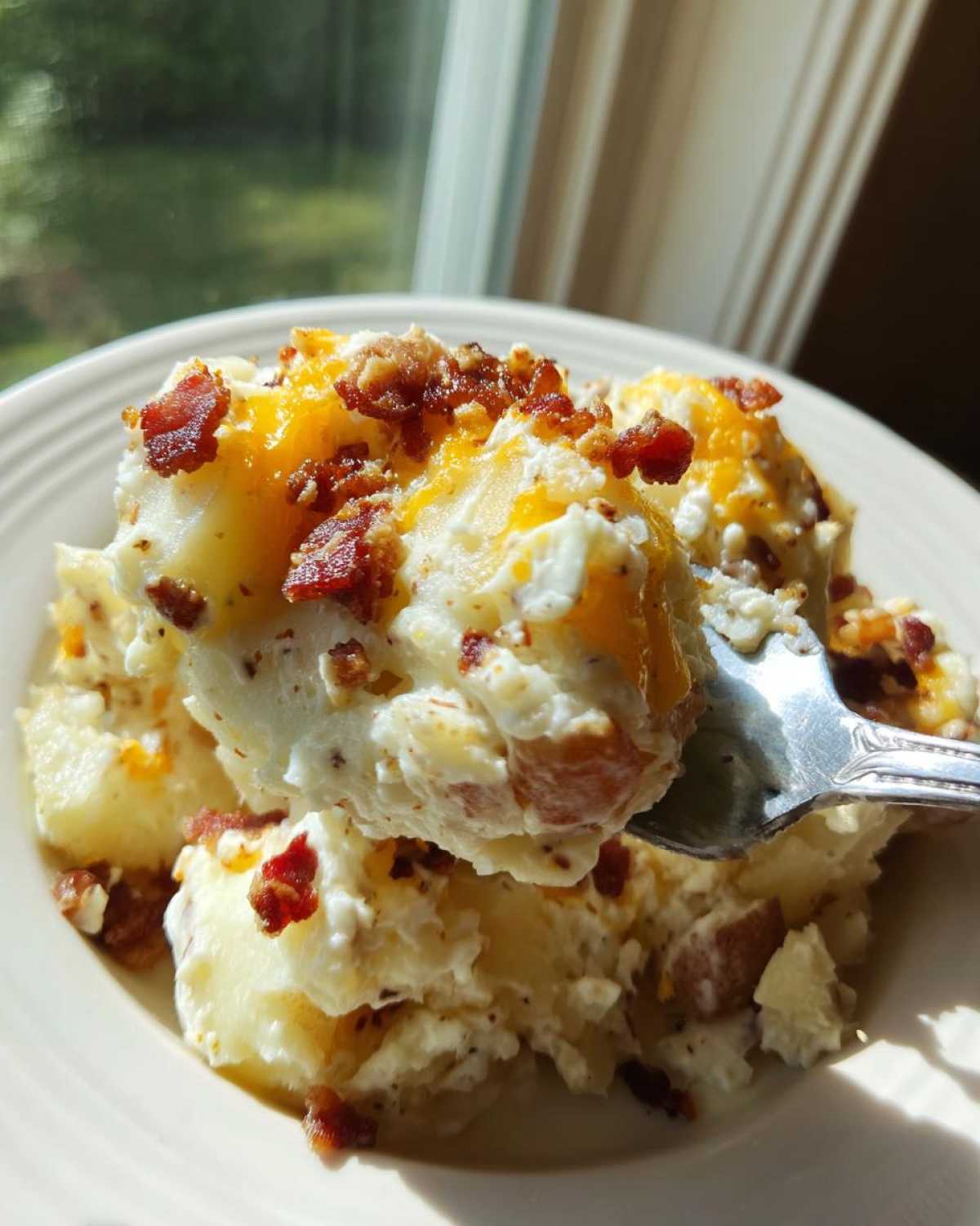 A fork lifting a creamy scoop of baked potato salad topped with melted cheese and bacon bits.