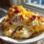 A spoonful of creamy baked potato salad topped with melted cheddar cheese and crispy bacon bits being lifted from a white plate.