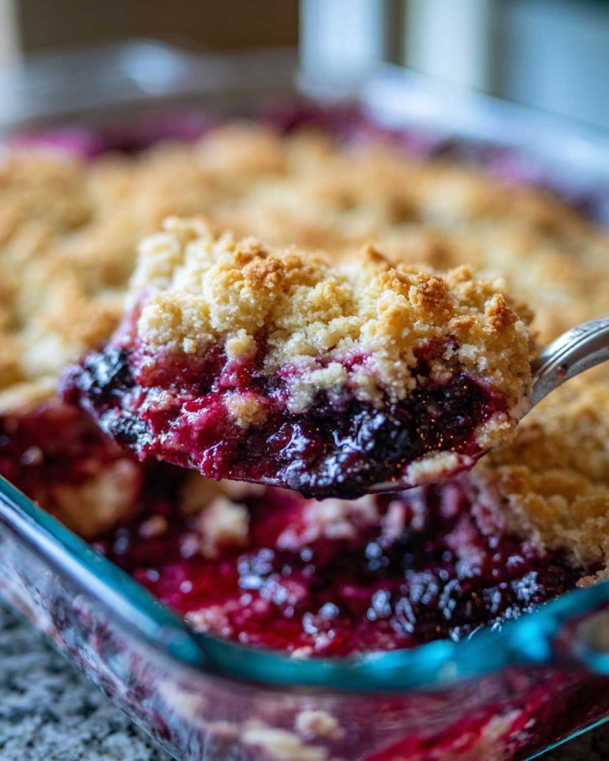 A spoonful of warm berry cobbler with a golden crumble topping being lifted from a glass baking dish.