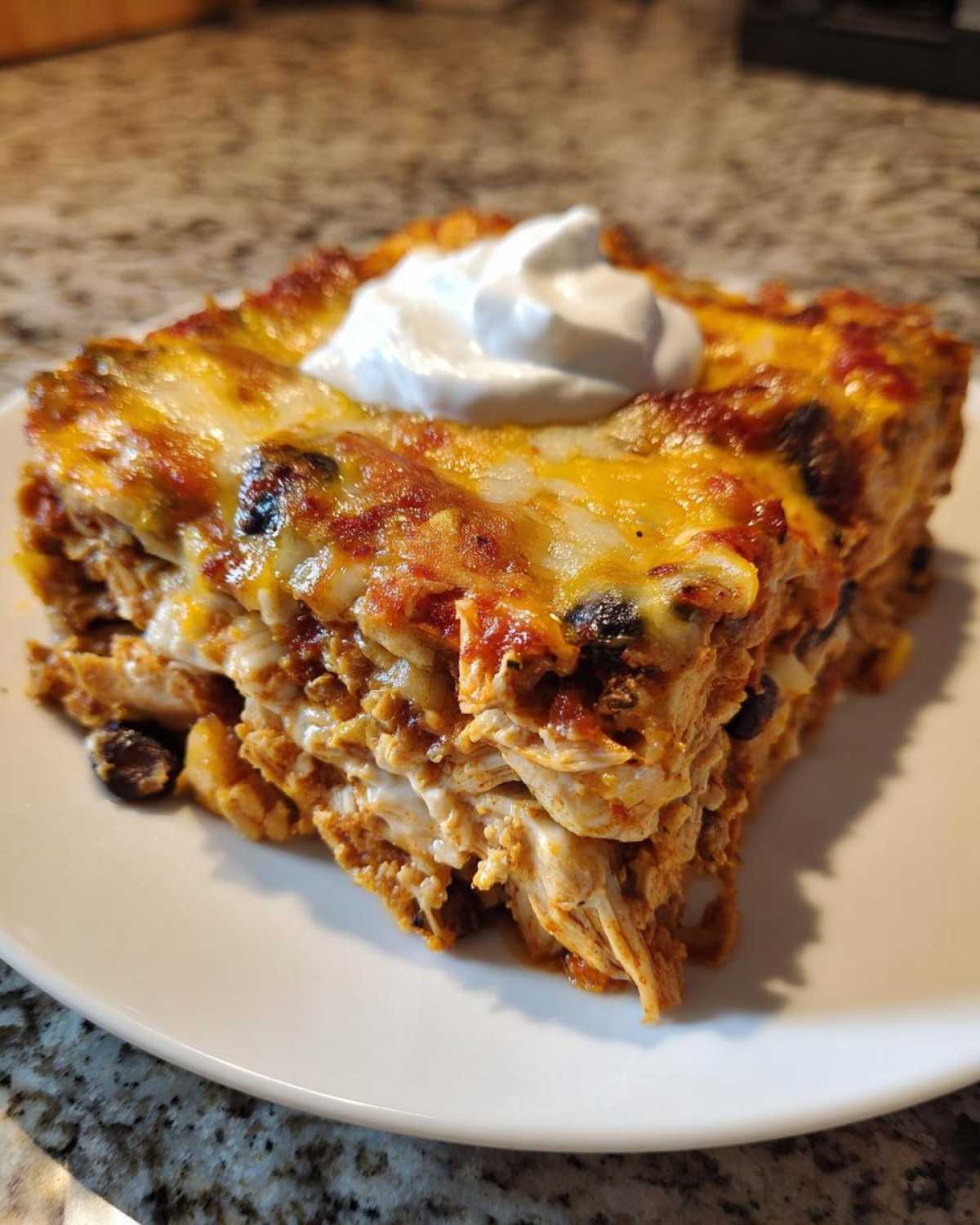 A square slice of cheesy, layered burrito bake topped with a dollop of sour cream on a white plate.