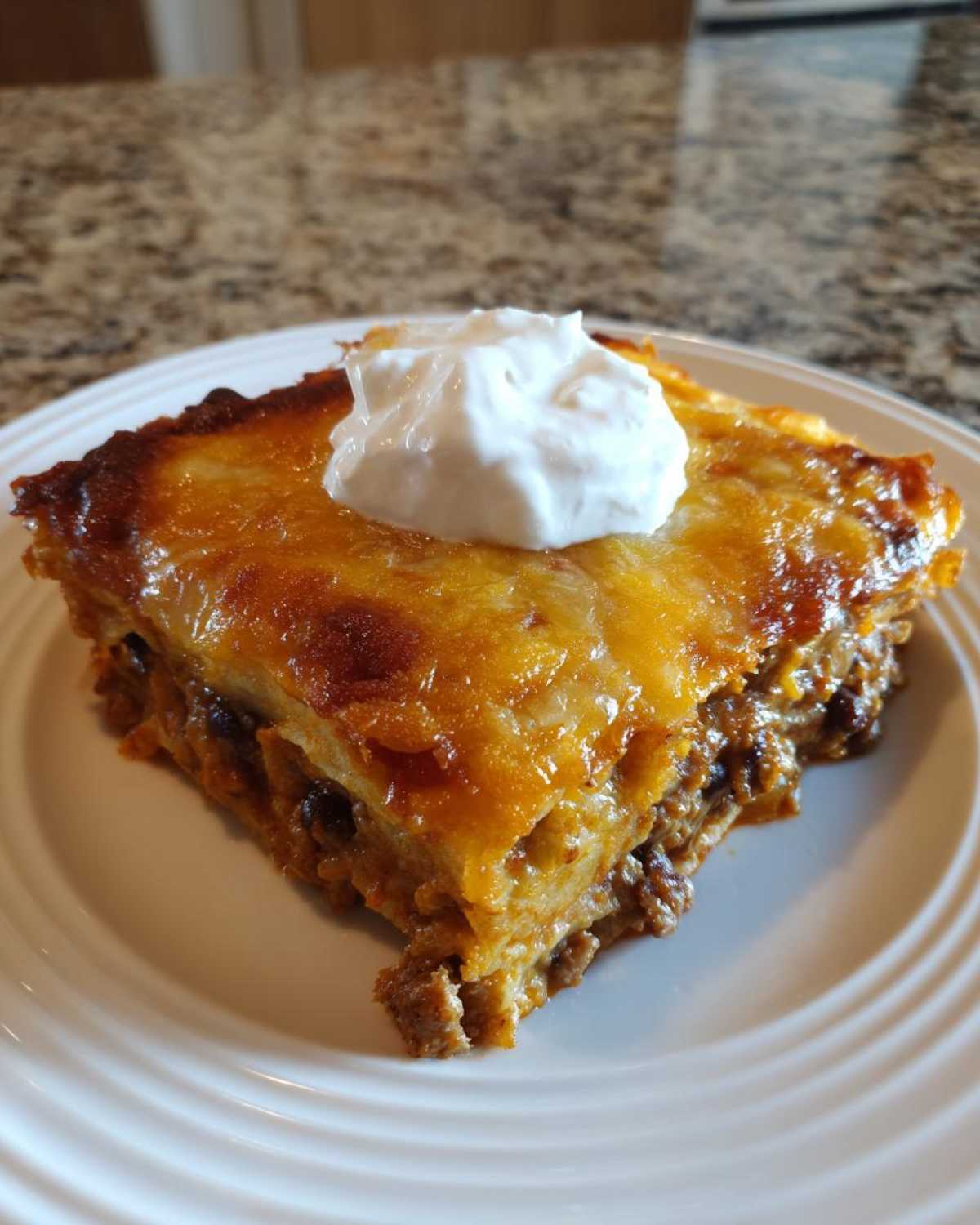 A square slice of cheesy, layered burrito bake topped with a dollop of sour cream on a white plate.