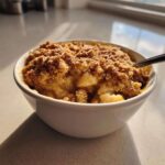 Close-up of a warm, single-serving apple crumble in a white bowl with a spoon, featuring a golden brown, crisp topping.