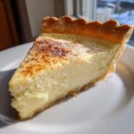 A close-up slice of creamy, yellow egg custard in a flaky crust, dusted with cinnamon.