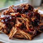 Close-up of tender, shredded Slow Cooker Pulled Pork generously coated in glossy dark BBQ sauce on a white plate.