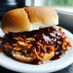 Close-up of a juicy Slow Cooker Pulled Pork sandwich smothered in barbecue sauce on a white plate.