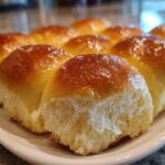 A close-up of freshly baked, soft dinner rolls with a shiny, golden-brown glaze on a white platter.