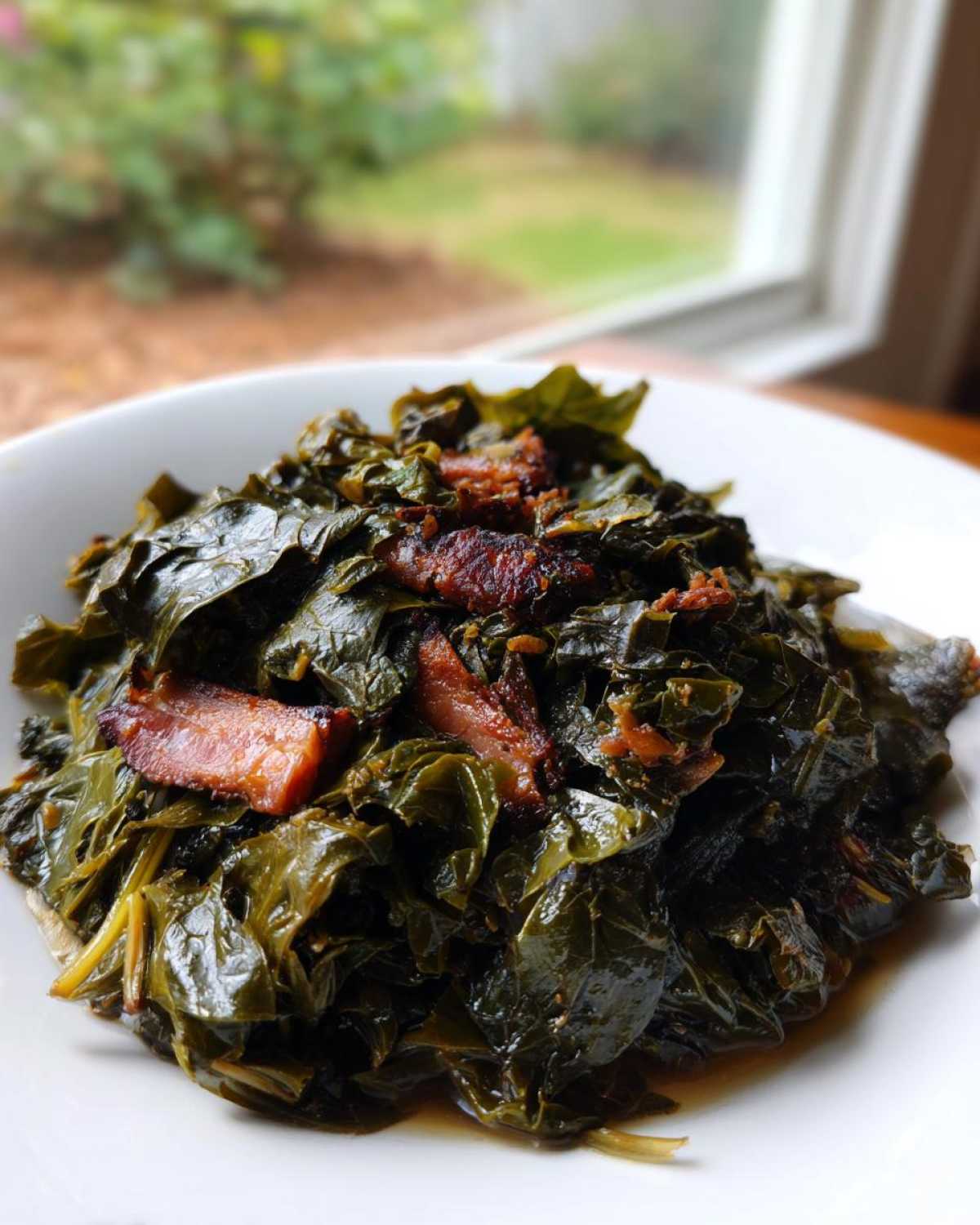 A close-up of tender, dark green collard greens served in a white bowl, topped with pieces of smoked ham.