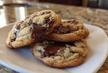 Three freshly baked sourdough chocolate chip cookies stacked on a white plate with gooey, melted chocolate.