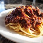 Close-up of spaghetti topped generously with rich, dark bolognese sauce on a white plate.