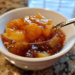 A spoonful of thick, chunky apple pie filling, rich with cinnamon, being lifted from a white bowl.