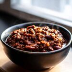 Close-up of a dark bowl filled with steaming, rich, dark red chili, highlighting the best chili recipe texture.