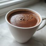 Close-up of a white mug filled with rich, dark hot cocoa mix, resting on a white marble surface near a window.