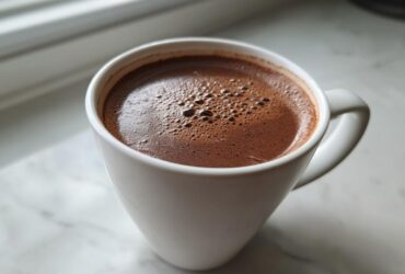 Close-up of a white mug filled with rich, dark hot cocoa mix, resting on a white marble surface near a window.