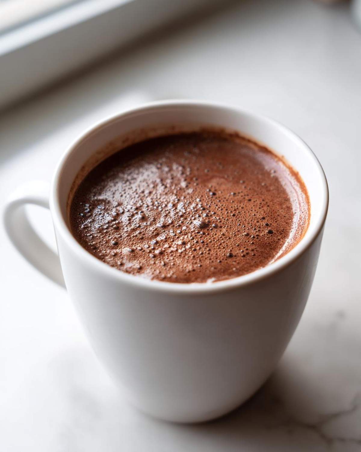 Close-up of a steaming white mug filled with rich, dark hot cocoa mix, showing frothy bubbles on top.